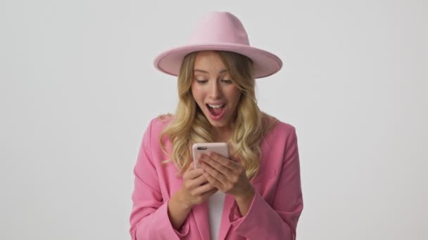 Surprised young blonde woman in pink jacket and hat becoming very happy and screaming loudly while using smartphone over gray background isolated - Filmagem, Vídeo