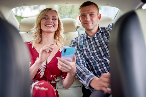 Image of smiling woman and man with phone in hands sitting in back seat of car . - Foto, Imagen