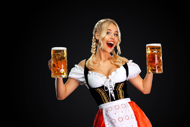 Happy sexy oktoberfest girl waitress, wearing a traditional Bavarian or german dirndl, serving two big beer mugs with drink isolated on black background. - Φωτογραφία, εικόνα