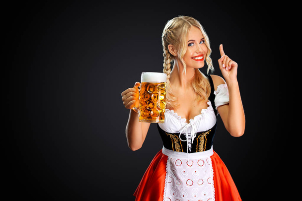 Smiling young sexy oktoberfest girl waitress, wearing a traditional Bavarian or german dirndl, serving big beer mug with drink isolated on black background. - Photo, image
