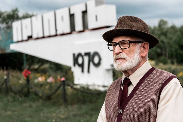 PRIPYAT, UKRAINE - AUGUST 15, 2019: senior bearded man in glasses standing near monument with pripyat letters - 写真・画像