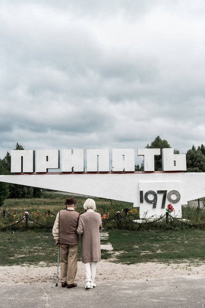 PRIPYAT, UCRANIA - 15 de agosto de 2019: vista trasera de los esposos jubilados de pie cerca del monumento con letras pripyat
 - Foto, Imagen