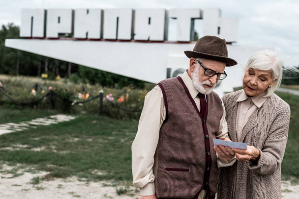 PRIPYAT, UKRAINE - AUGUST 15, 2019: retired couple looking at photo near monument with pripyat letters  - Zdjęcie, obraz