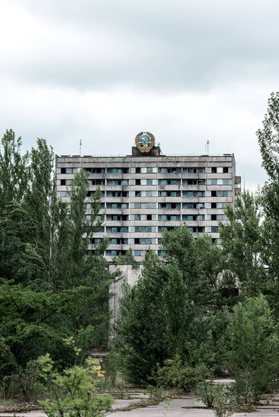 PRIPYAT, UKRAINE - AUGUST 15, 2019: green trees near building against sky with clouds in chernobyl  - Fotó, kép