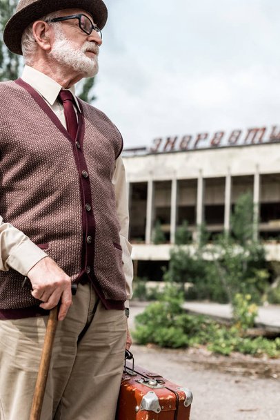 PRIPYAT, UKRAINE - AUGUST 15, 2019: senior man holding suitcase near building with energetic lettering in chernobyl  - Фото, изображение