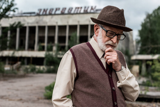 PRIPYAT, UKRAINE - AUGUST 15, 2019: pensive senior man standing near building with energetic lettering in chernobyl  - Photo, image