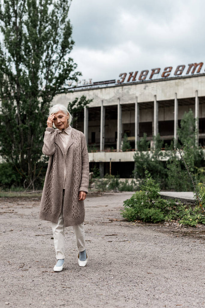 PRIPYAT, UKRAINE - AUGUST 15, 2019: senior woman walking near building with energetic lettering in chernobyl  - Fotoğraf, Görsel