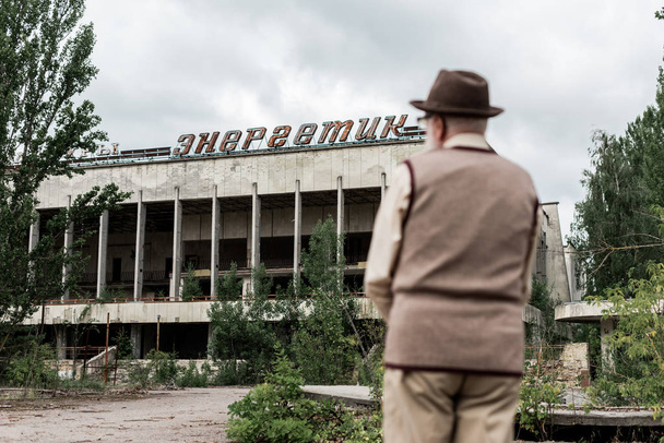 PRIPYAT, UKRAINE - AUGUST 15, 2019: back view of retired man standing near building with energetic lettering in chernobyl  - Φωτογραφία, εικόνα