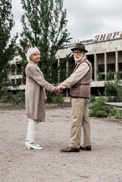 PRIPYAT, UKRAINE - AUGUST 15, 2019: senior couple holding hands near building with energetic lettering in chernobyl  - Foto, imagen