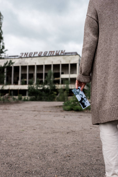 PRIPYAT, UKRAINE - AUGUST 15, 2019: cropped view of retired woman holding black and white photo near building in chernobyl  - Photo, Image