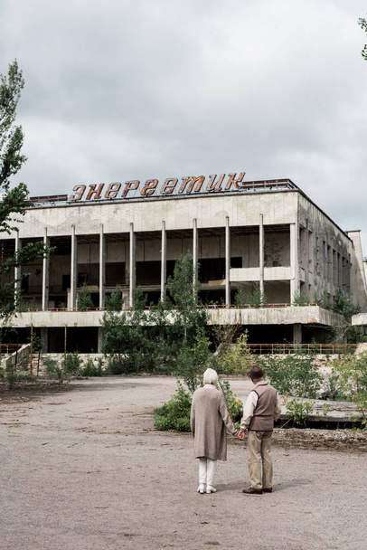 PRIPYAT, UKRAINE - AUGUST 15, 2019: back view of retired couple holding hands near building with energetic lettering in chernobyl  - Valokuva, kuva