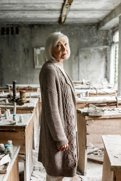 selective focus of frustrated retired woman standing in dirty abandoned classroom  - Photo, Image