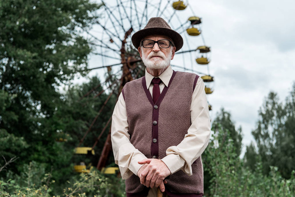 PRIPYAT, UKRAINE - AUGUST 15, 2019: bearded retired man in hat and glasses standing with walking cane in amusement park with ferris wheel  - Foto, Imagen