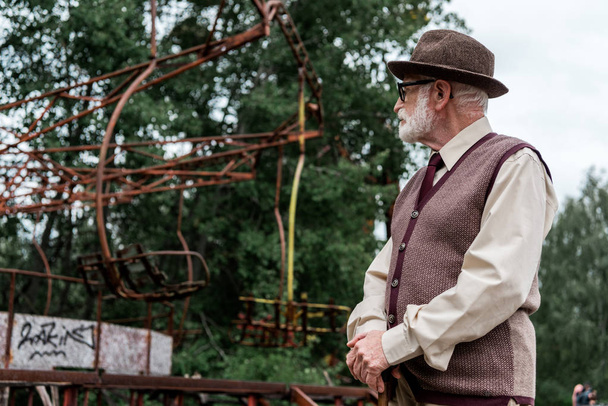 PRIPYAT, UKRAINE - AUGUST 15, 2019: bearded retired man in hat and glasses standing with walking cane and looking at damaged carousel  - Photo, Image