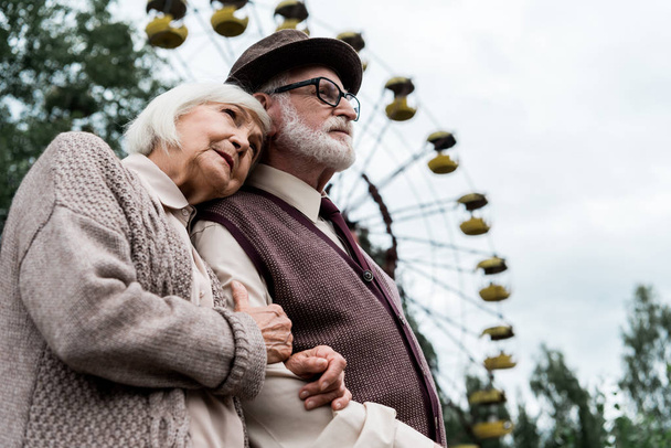 PRIPYAT, UKRAINE - AUGUST 15, 2019: low angle view of senior woman hugging husband near ferris wheel in amusement park - Photo, Image