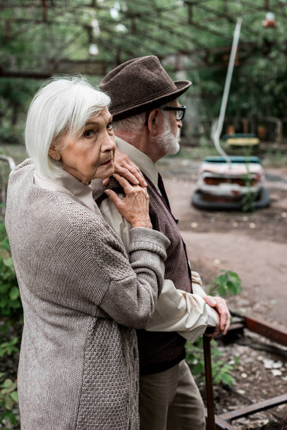 PRIPYAT, UKRAINE - AUGUST 15, 2019: senior man in hat standing with retired wife near amusement park in chernobyl  - Photo, image