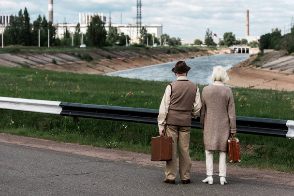 PRIPYAT, UKRAINE - AUGUST 15, 2019: back view of senior couple with baggage near chernobyl nuclear power plant  - Photo, Image