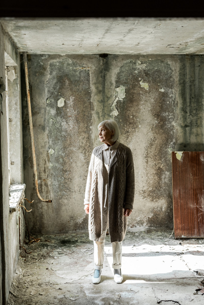 ηλικιωμένη γυναίκα με γκρίζα μαλλιά στέκεται σε άδειο και βρώμικο δωμάτιο  - Φωτογραφία, εικόνα