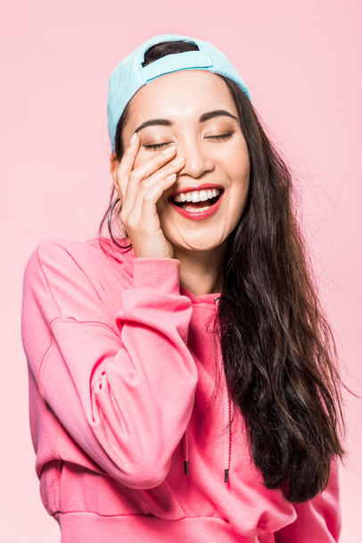 attractive asian woman with closed eyes in pink pullover and cap smiling isolated on pink  - 写真・画像