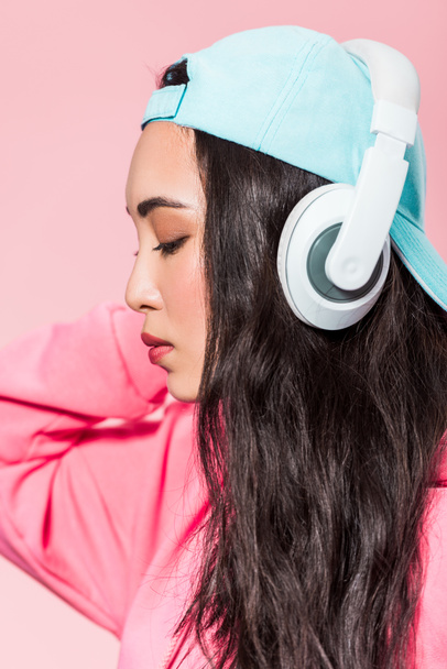 attractive asian woman in pullover and cap listening music isolated on pink  - Foto, Imagem