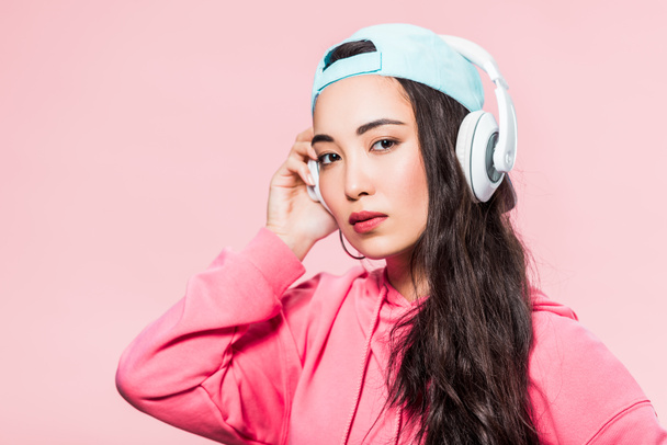 attractive asian woman in pullover and cap listening music isolated on pink  - Foto, imagen