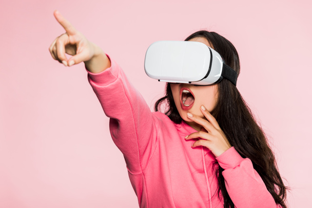 shocked woman in pink pullover with virtual reality headset pointing with finger isolated on pink  - Fotografie, Obrázek