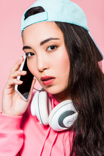 attractive asian woman in pullover and cap with headphones talking on smartphone isolated on pink  - Foto, Bild
