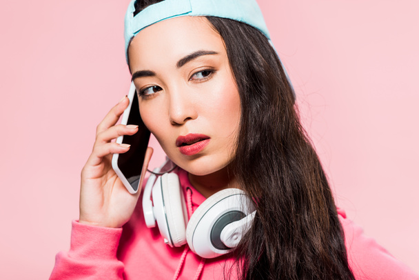 attractive asian woman in pullover and cap with headphones talking on smartphone isolated on pink  - Φωτογραφία, εικόνα