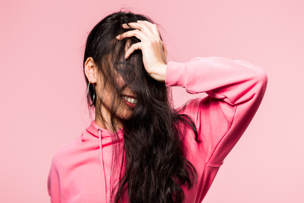 attractive asian woman in pink pullover smiling and obscuring face isolated on pink  - Фото, изображение