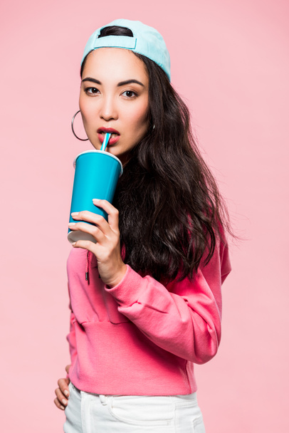 attractive asian woman in pullover and cap drinking from plastic cup isolated on pink  - Fotó, kép