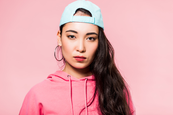 attractive asian woman in pink pullover and cap looking at camera isolated on pink  - Foto, imagen