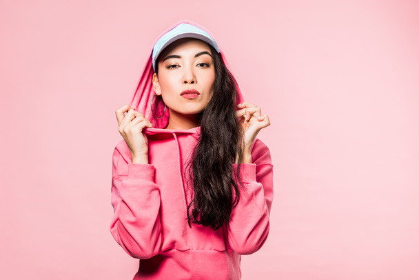 attractive asian woman in pink pullover and cap looking at camera isolated on pink  - Photo, Image