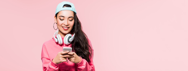 panoramic shot of asian woman in pullover and cap with headphones holding smartphone isolated on pink  - Valokuva, kuva