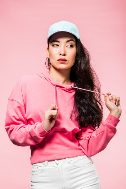 attractive and pensive asian woman in pink pullover and cap looking away isolated on pink  - Фото, зображення