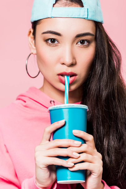 attractive asian woman in pullover and cap drinking from plastic cup isolated on pink  - Photo, Image