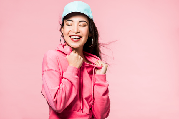 attractive asian woman with closed eyes in pink pullover and cap smiling isolated on pink  - Fotografie, Obrázek