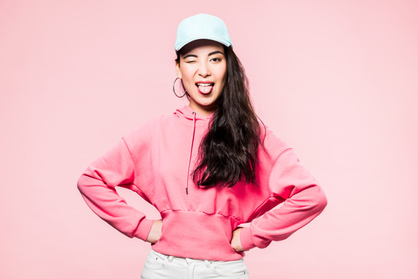 attractive asian woman in pink pullover and cap winking and sticking out tongue isolated on pink  - Foto, imagen