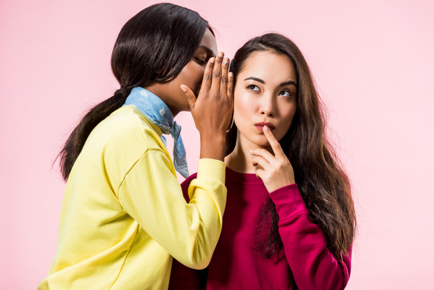 african american woman telling secret to her asian friend isolated on pink  - 写真・画像