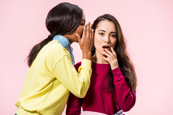african american woman telling secret to her shocked asian friend isolated on pink  - Foto, Imagen