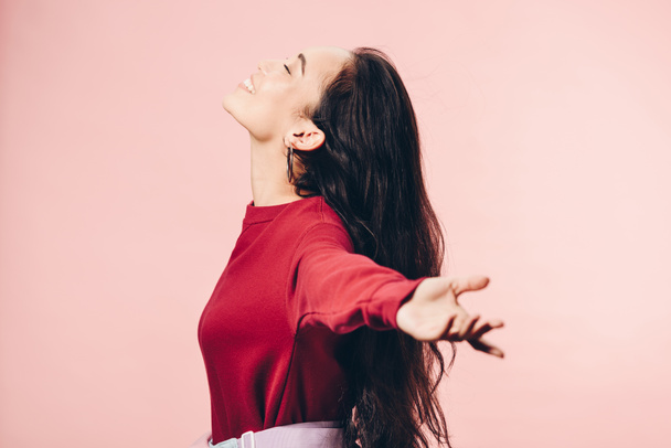 side view of attractive asian woman with outstretched hands in red sweater smiling isolated on pink  - Foto, Imagem