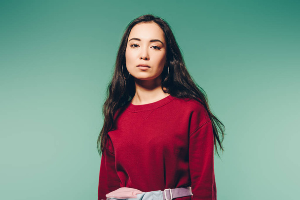 attractive asian woman in red sweater looking at camera isolated on green   - Фото, изображение