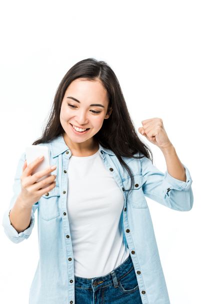 ελκυστική Ασιάτισσα γυναίκα με denim πουκάμισο δείχνει ναι χειρονομία και κρατώντας smartphone απομονώνονται σε λευκό  - Φωτογραφία, εικόνα