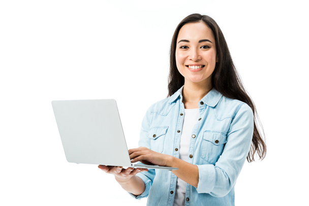 attractive asian woman in denim shirt smiling and holding laptop isolated on white  - 写真・画像