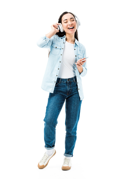 attractive and smiling asian woman in denim shirt listening music and holding smartphone isolated on white  - Foto, Bild