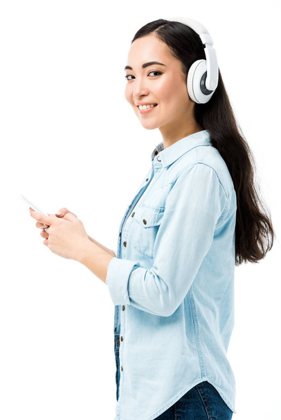 atraktivní a usměvavá asijská žena v džínové košili poslech hudby a držení smartphone izolované na bílém  - Fotografie, Obrázek