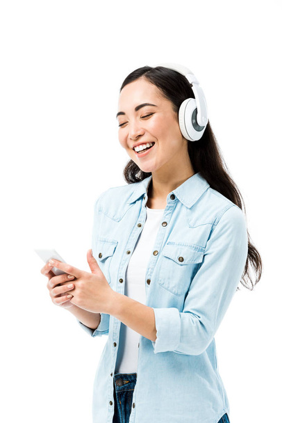 attraktive und lächelnde asiatische Frau in Jeanshemd, die Musik hört und ihr Smartphone isoliert auf weiß hält  - Foto, Bild