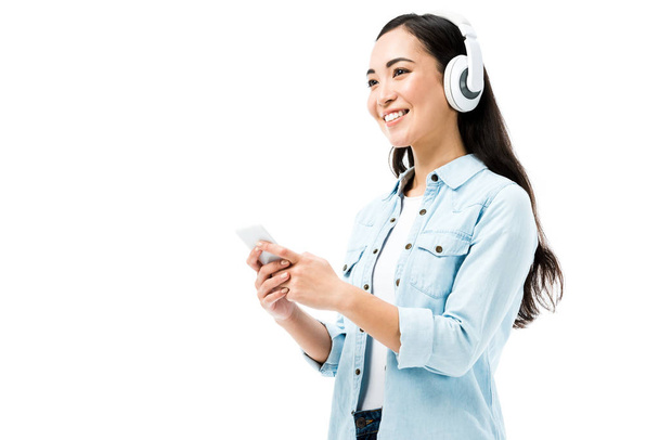 attractive and smiling asian woman in denim shirt listening music and holding smartphone isolated on white  - Fotó, kép