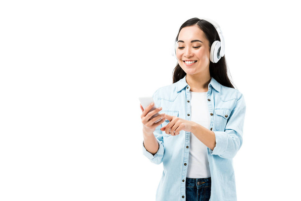 atrakcyjna i uśmiechnięta Azjatka w dżinsowej koszuli słuchająca muzyki i trzymająca smartfona odizolowanego na biało  - Zdjęcie, obraz