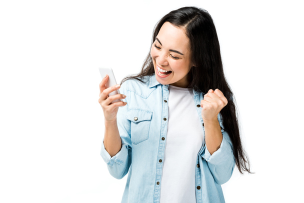 atraktivní a usměvavá asijská žena v džínové košili ukazující ano gesto a držení smartphone izolované na bílém  - Fotografie, Obrázek