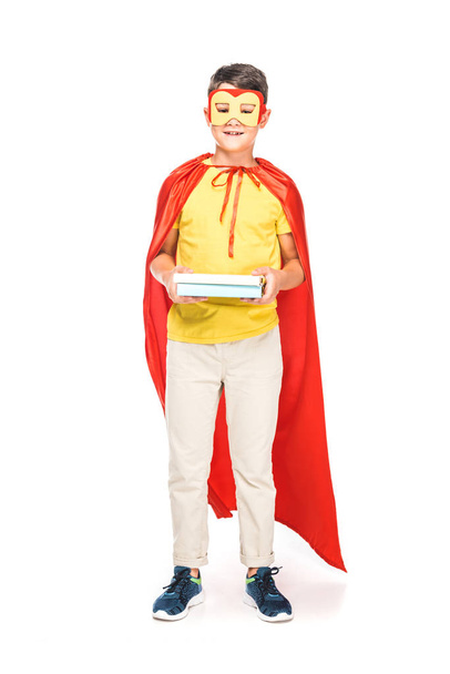 full length view of kid in mask and hero cloak holding books on white - Фото, изображение